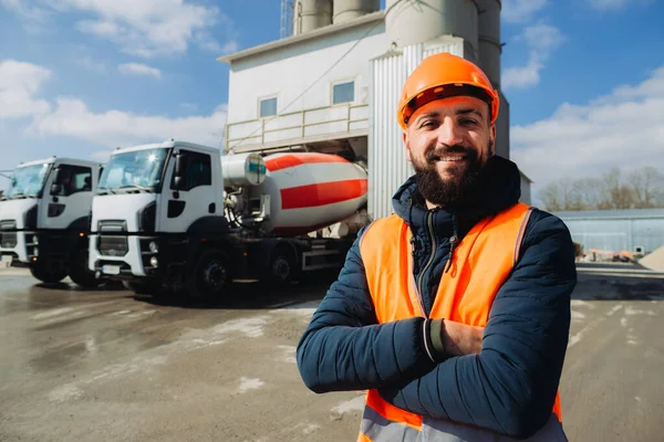 Çimento kamyonları ve beton fabrikasının önünde kolları çaprazlanmış gülümseyen bir inşaat işçisinin portresi.