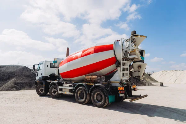 Güneşli bir günde çimento fabrikasında park etmiş kırmızı ve beyaz beton karıştırıcı kamyon. Arka planda kum ve çakıl yığınları var.