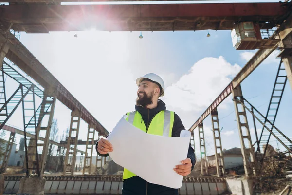 Güvenlik yeleği ve miğfer takan sakallı mühendis planlar tutuyor ve beton üretim tesisine gülümsüyor.