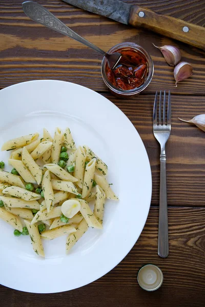 Pasta with pesto sauce, green peas, garlic and dill on a white plate on a wooden table. Near are a fork,a knife and sun-dried tomatoes in a jar. Vegetarian cuisine. The concept of healthy eating