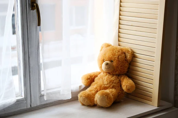 A cute little Teddy bear toy sits on a brown wooden and white plastic windowsill with a copy of the text space on the window glass. Beautiful lonely bear on the window. The concept of childhood.