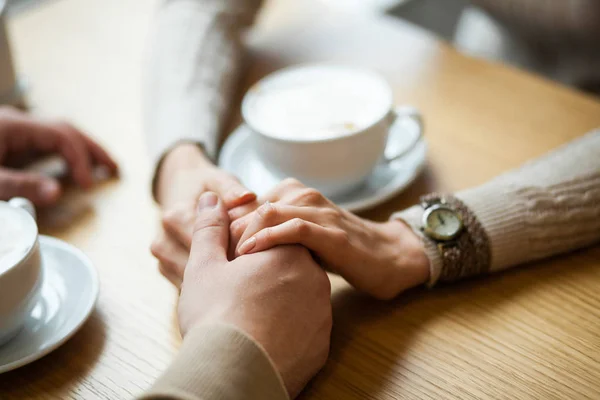Couple hold hands drinking coffee in cafe, close up of lovers arms on background of wooden table. Breakfast or lunch in restaurant. Engagement in cafe, boyfriend tightly holding his girlfriend hand