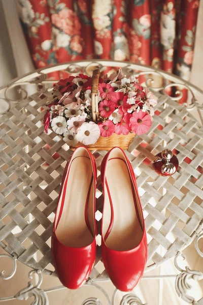 Wedding red designer bride shoes, perfume bottle and decorative flowers in a basket on the coffee table. Women\'s new luxury modern fashion shoes made of shiny patent leather. Wedding theme