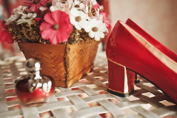 Wedding red designer bride shoes, perfume bottle and decorative flowers in a basket on the coffee table. Women\'s new luxury modern fashion shoes made of shiny patent leather. Blurred background