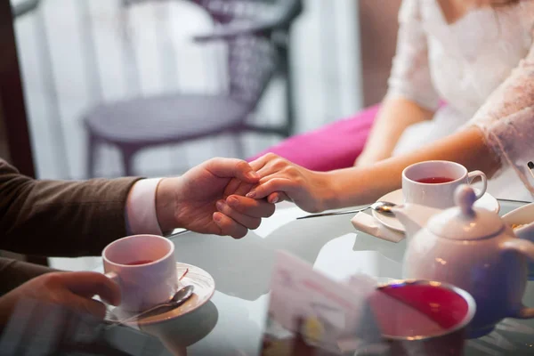 Loving couple holding hands, drinking tea in a cafe at the glass table. Breakfast or lunch at the restaurant. Engagement, anniversary and wedding day. The guy firmly holding his girlfriend\'s hand.