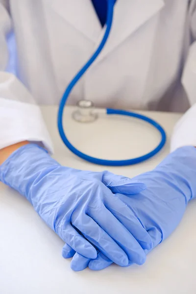 Hands of the attending physician in medical rubber gloves on the background of a white table, close-up. Doctor in scrubs with a stethoscope. The concept of health, pharmacy and medicine.