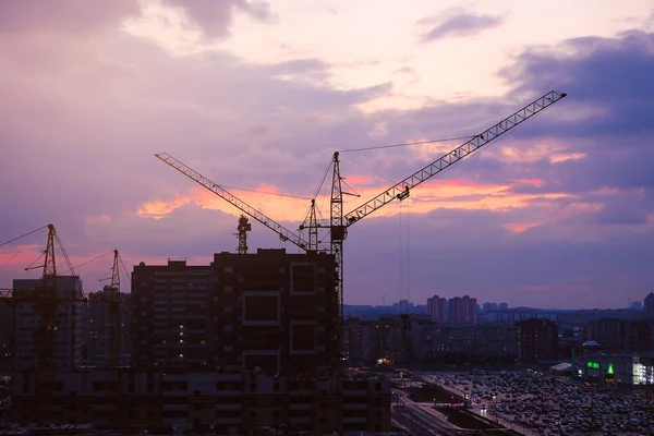 Tower crane, new buildings, Parking and cars in the distance during the sunset of the city, against the sky. Beautiful night city. Residential area with houses under construction.
