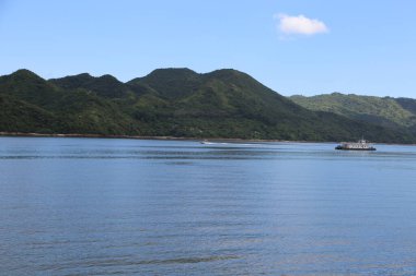 Denizde küçük bir yolcu feribotu, arkasında yeşil dağlar var. Su durgun ve mavidir. Doğanın güzelliğinin huzurlu bir sahnesini yaratır.