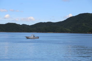 Bir dağın önünde suda yüzen bir tekne var. Tekne beyaz ve üzerinde bir adam var.
