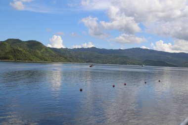 Geniş deniz su yüzeyinin panoramik bir görüntüsü, uzak dağlar ve arka planda mavi gökyüzü. Önünde yoğun yeşil bitki örtüsüyle kaplı bir ada var. Üzerinde yüzen küçük bir tekne var..