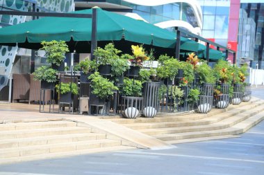 basamaklarda modern yeşil bitkilerle dolu bir sokak kenarı kafesi, her masada tentelerin altında duran büyük koyu gri metal top saksıları, lüks alışveriş ve ofis binaları yakınlarındaki bir şehir meydanında..