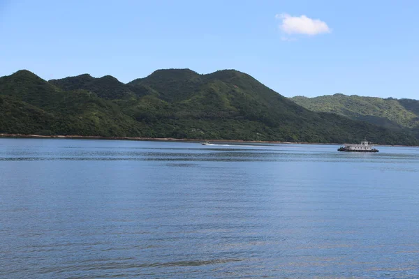 Denizde küçük bir yolcu feribotu, arkasında yeşil dağlar var. Su durgun ve mavidir. Doğanın güzelliğinin huzurlu bir sahnesini yaratır.