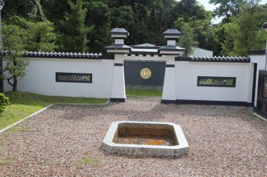 Siyah camlı beyaz bir duvar ve üzerinde altın rengi bir logo var. Etrafı çakıl taşlarıyla çevrili. Önünde açık bir ateş çukuru var. Arka planda, Çin mimarisi tarzında yemyeşil ağaçlar var..