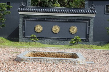 Çin tarzındaki villanın gri duvarı, üzerinde üç altın dairesel oyma ile süslenmiş ve önünde çakıl taşlarıyla kaplı açık bir bahçe var.