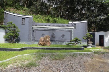 Üzerinde yeşil çimenler ve önünde iki bonsai ağacı olan gri bir duvar. Ayrıca bir tarafta taş oymalar için de bir alan var..