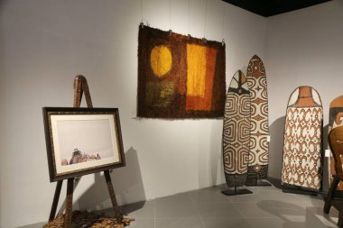an art gallery featuring african tribal patterns, wall hangings, and an ironing board as artwork on display. the room is well-lit with natural light. a large framed painting hangs above the easel.