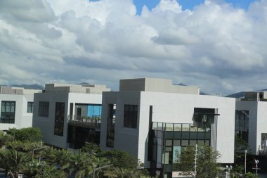 a view of three modern buildings in the city center with white facades and black windows, overlooking green parkland with tropical trees and a blue sky with clouds