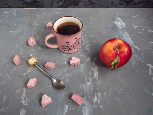 Red apple, a cup of coffee and pink lump sugar, a teaspoon on a gray background