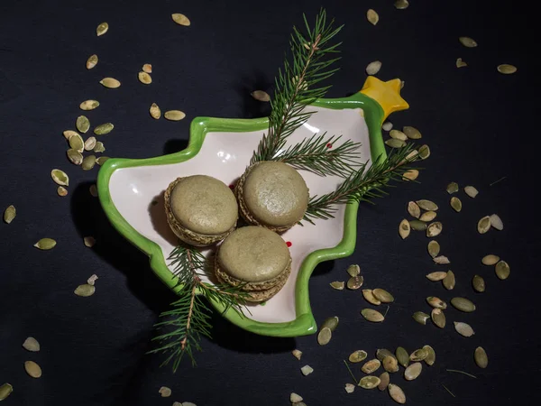Macarons à la pistache sur une assiette du Nouvel An sur un fond sombre, une dispersion de graines de citrouille. Branche d'épinette. Gros plan . — Photo