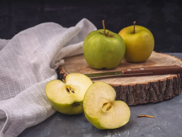 Two green golden apples and two halves on a wooden platter and gray background, cotton white napkin, kitchen knife. Close-up.