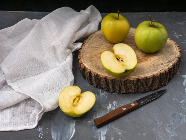 Two green golden apples and two halves on a wooden platter and gray background, cotton white napkin, kitchen knife. Close-up.
