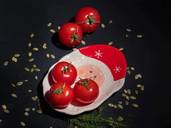 Red tomatoes with leaves on a decorative Christmas plate. Pumpkin seeds are scattered on a black background