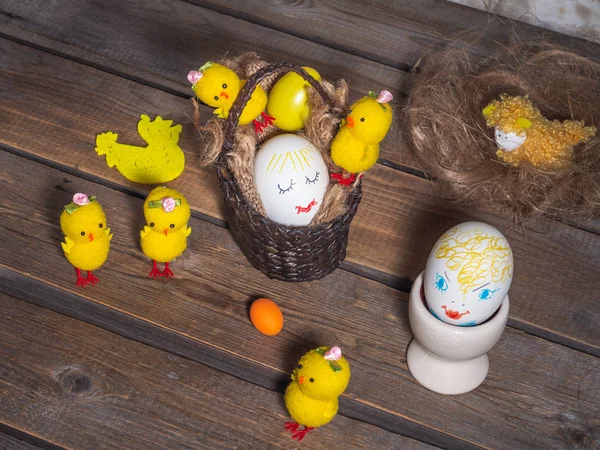 Easter fun picture with painted faces on the eggs, toy chickens are located on a wooden old background. Prefabricated basket and egg holder.