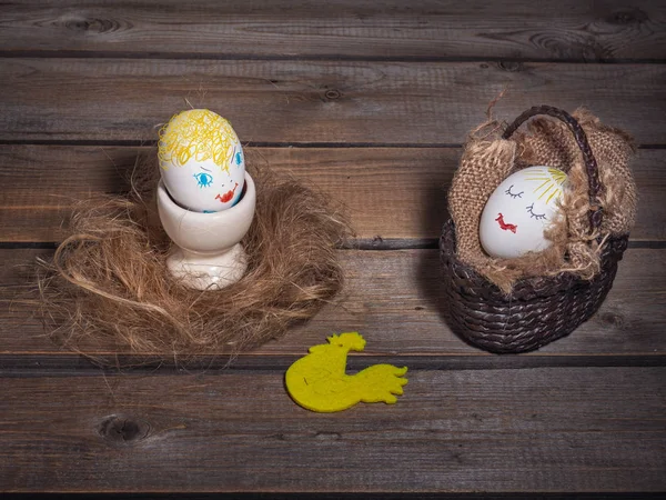 Fun picture with eggs with painted faces. One is in a wicker basket, and the other is on an egg holder. Old wooden background, cockerel figurine.