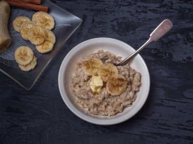 Healthy breakfast of oatmeal, sprinkled with cinnamon and banana slices. Teaspoon and plates on a wooden gray background. View from above.