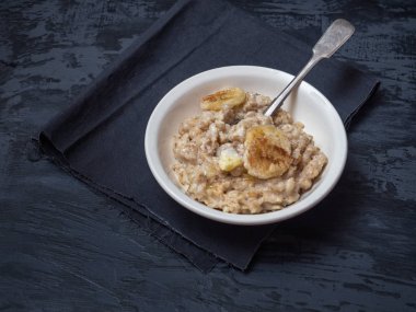 Yulaf lapası ve bir kaşık, tarçın ve muz daireler iã§in derin bir seramik tabak. Tabağın altında koyu bir peçete var. Koyu ahşap arka plan üzerinde çekim yakın.