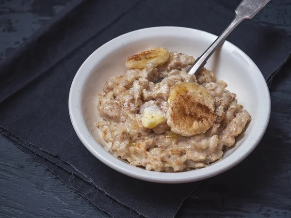 Deep ceramic plate with porridge, sprinkled with cinnamon and banana circles. There is a dark cotton napkin under the plate. Close-up shot on a dark wooden background.