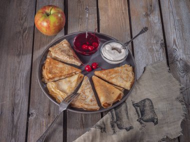 Pancakes rolled by a triangle on a large gray plate, a number of small ice-cream bowls with sour cream and canned cherries, a large apple, tableware on the plate and a linen napkin near the plate.