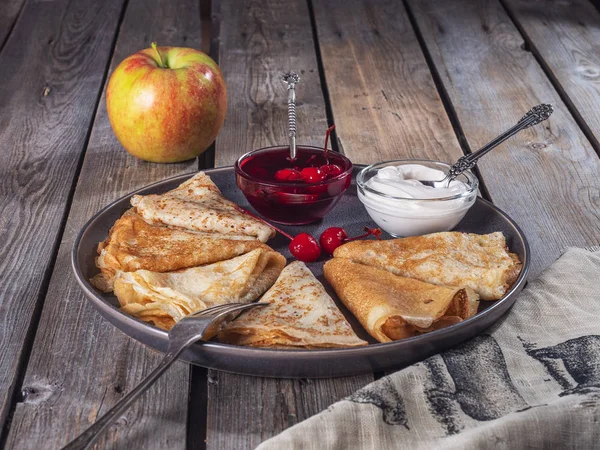 Pancakes rolled by a triangle on a large gray plate, next to small ice-cream bowls with sour cream and canned cherries, a large apple in the background, tableware for the plate and a linen napkin near