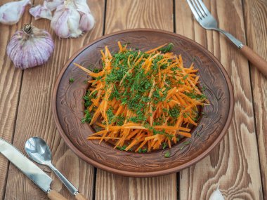 Kahverengi seramik plaka içinde dereotu ve bitkisel yağ ile taze havuç salatası. Arka planda taze sarımsak. Tahta masasında çatal bıçak takımı,