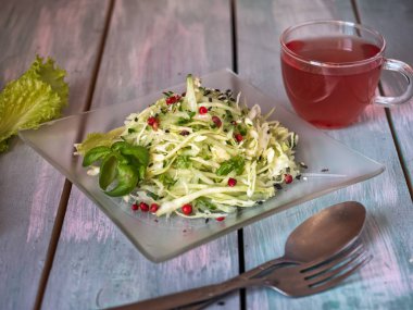 Bir cam dekoratif plaka üzerinde renkli biber bezelye ile taze lahana ve salatalık bir salata, yaprak marul yaprakları plaka yanında yatıyor. Tahta tepside çatal bıçak takımı.