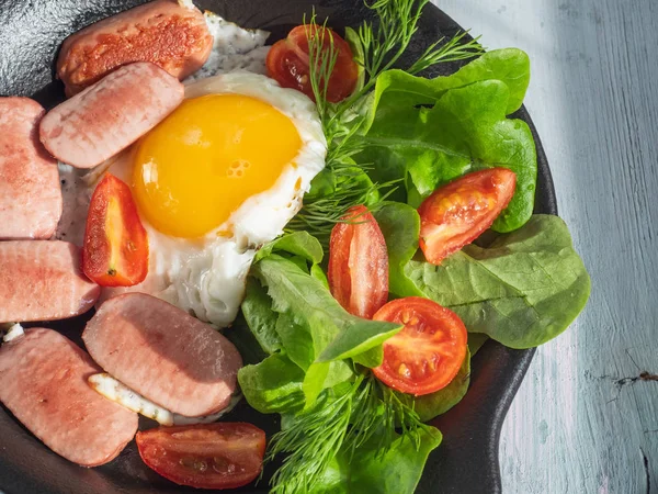 Breakfast of fried eggs with sausages, cherry tomatoes and leafy lettuce on a black round pan. Serve on a wooden plank tray with bread rolls as a reference fork.