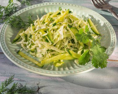 Yakın mesafeden alınan bir tahta masada yuvarlak marul tabağı üzerinde baharatlı otlar ile taze katusta ve salatalık ile hafif salata