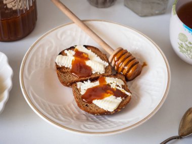 Beyaz seramik tabakta tereyağı ve bal ile tatlı sandviçler. Cam kavanozda bal, bal için kaşık.
