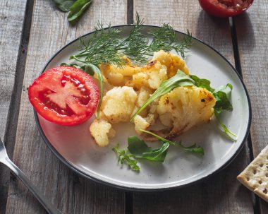 ahşap bir masa üzerinde yuvarlak bir seramik tabakta arugula ve domates ile kızarmış karnabahar