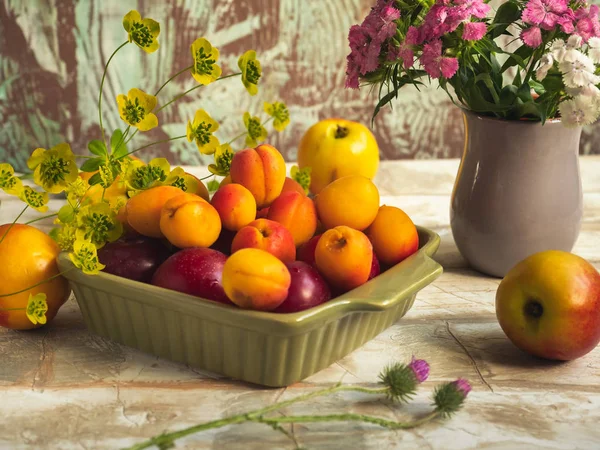 Fruit set of ripe fruits. Apricots and nectarines in a green plate. A small bouquet of carnations in a gray vase.