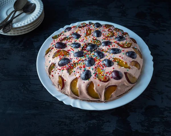 Homemade cake with fruit and sour cream on a white plate on a black table, in the background of a saucer with spoons — Stock Photo, Image