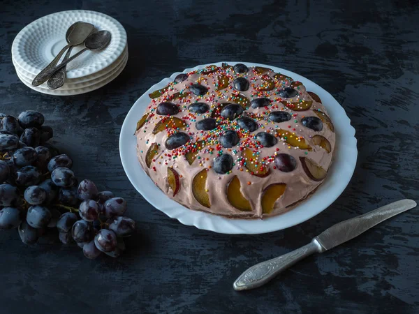 Homemade cake. Cake with fruit and berries on a dark table, a branch of grapes and a stack of saucers for distributing cake. — Stock Photo, Image
