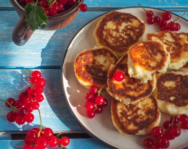 Kırmızı frenk üzümü sprigs ile yuvarlak bir tabak üzerinde ev yapımı cheesecakes, mavi ahşap tepsi üzerinde üst görünümü