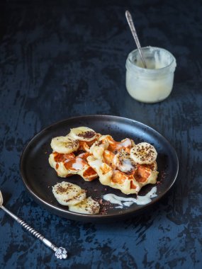 Siyah bir tabakta muz kremalı waffle, cam bir kavanozda bal ve siyah bir arka planda kaşık.