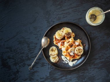 Siyah bir tabakta muz ve bal ile waffle ve bal kavanozu ve yukarıdan siyah arka plan manzaralı bir kaşık.