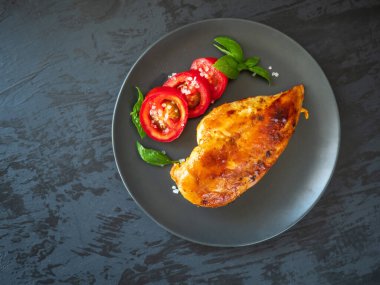 Fırında tavuk fileto ve fesleğen yapraklı dilimlenmiş kırmızı domatesler gri bir fonda gri bir tabakta.