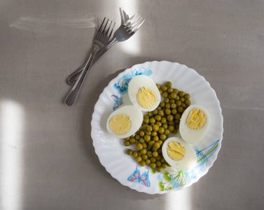 Hafif kahvaltıda haşlanmış yumurta ve hafif tabakta yeşil bezelye, gri arka planda çatallar.