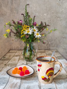 Hafif öğle yemeği, büyük seramik bardakta çay, içinde horoz resmi ve marmelatlı bir fincan çay, kırsal mutfak masasındaki cam sürahide bir buket yabani çiçek.