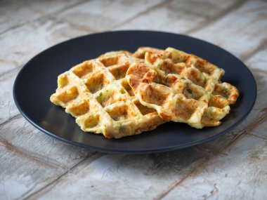 Kapalı bir tabakta, ahşap dekoratif bir masanın üzerinde duran yeşil ve peynirli kabaklı waffle.