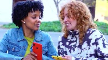 Two smiling multiracial women sitting at outdoor table using smartphones, modern lifestyle, leisure, friendship, digital technology, online communication, social connection, mobile app, young people